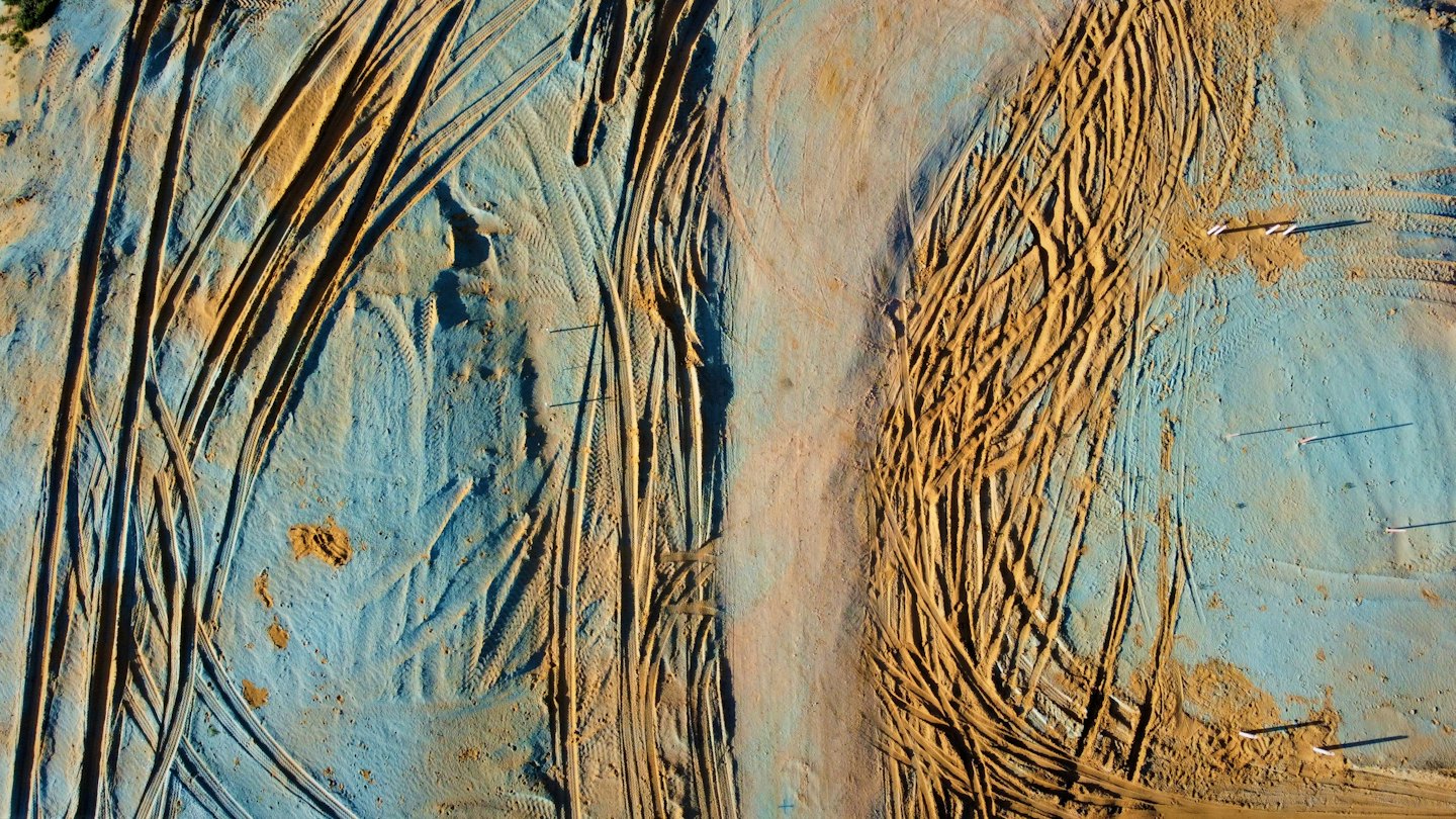Tracce pesanti di pneumatici attraversano un cantiere con picchetti di rilevamento.